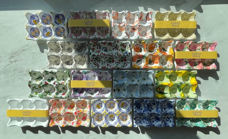 Peut inclure: Une collection de bo&icirc;tes &agrave; &oelig;ufs rectangulaires en c&eacute;ramique, chacune dot&eacute;e de six alv&eacute;oles circulaires. Les bo&icirc;tes pr&eacute;sentent divers motifs color&eacute;s, notamment floraux, fruitiers et g&eacute;om&eacute;triques. Plusieurs sont entour&eacute;es d'une bande de papier jaune marqu&eacute;e CASITA FELIZ.