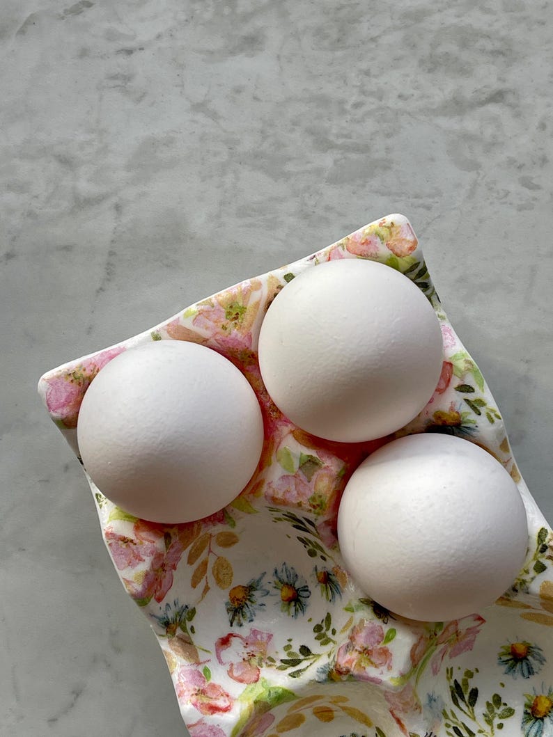 Peut inclure: Un coquetier en c&eacute;ramique avec une base blanche orn&eacute;e d'un motif floral color&eacute; compos&eacute; de fleurs roses, de c&oelig;urs jaunes et de feuilles vertes. Trois &oelig;ufs de poule blancs, lisses et de forme ovale sont nich&eacute;s dans les compartiments moul&eacute;s.