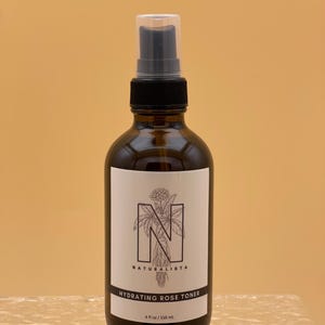 Könnte beinhalten: Eine braune Glasflasche mit schwarzem Sprühkopf und weißem Etikett. Das Etikett zeigt eine Blumenillustration und den Text "NATURALISTA HYDRATING ROSE TONER". Die Flasche enthält 118 ml Produkt.