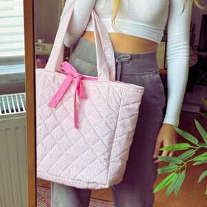 May include: A pink quilted tote bag with a white gingham pattern and a pink bow. The bag has a large capacity and is perfect for carrying everyday essentials.