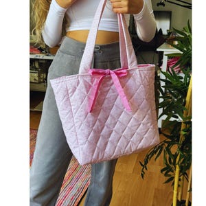 May include: A pink and white quilted tote bag with a pink gingham pattern and a pink bow. The bag has a quilted diamond pattern and is made of a soft, durable fabric.