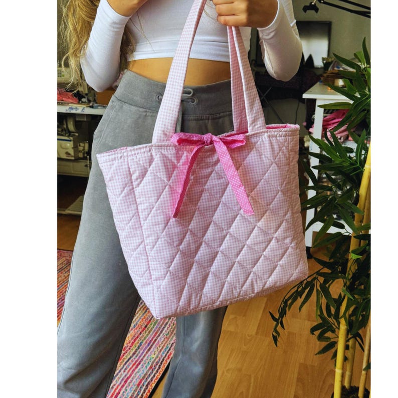 May include: A pink and white quilted tote bag with a pink gingham pattern and a pink bow. The bag has a quilted diamond pattern and is made of a soft, durable fabric.