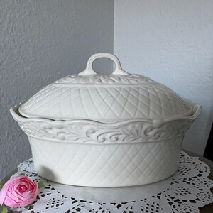 May include: White ceramic oval serving dish with a lid and a decorative handle. The dish has a woven pattern and a raised floral design around the rim.