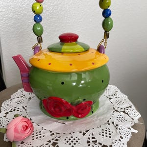 May include: A green ceramic teapot with a yellow lid and red roses on the side. The teapot has a colorful beaded handle and a pink spout.