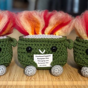 Peut inclure: Trois figurines de poubelles faites au crochet, avec des flammes de fil rouge, orange et jaune. Les poubelles vertes ont des yeux noirs, des roues grises et une étiquette blanche avec du texte. Le texte sur l'étiquette indique "Emotional Support Dumpster Fire."