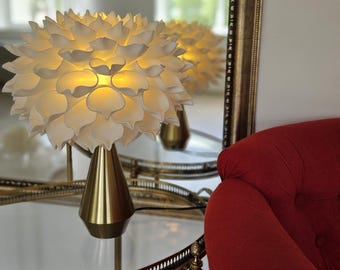May include: A table lamp featuring a white, multi-layered, petal-shaped shade that emits a warm, soft glow. The lamp has a smooth, conical, metallic gold base that tapers toward the top, providing a stable foundation for the floral-inspired shade.