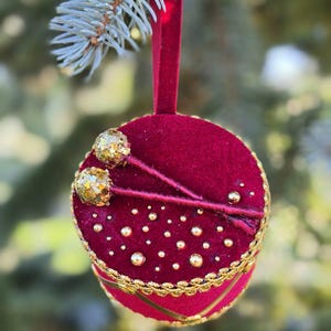 Op de afbeelding: Een rond ornament van dieprood fluweel met een gouden gevlochten rand langs de zijkanten. De bovenzijde is versierd met twee goudkleurige glitter-trommelstokjes en een strooiing van kleine, ronde gouden kralen in een stippenpatroon.