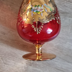 Può includere: Un calice di vetro rosso con bordo e base dorati. Il calice è decorato con un motivo floreale dorato e un piccolo gruppo di fiori dipinti.