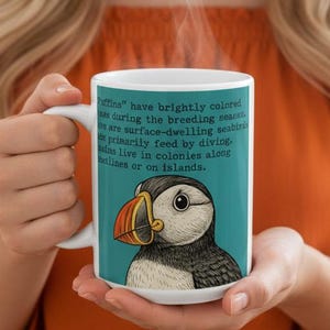 May include: White ceramic mug featuring a colorful illustration of a puffin bird with a large orange and yellow beak. The mug has a teal background with text describing puffins. The mug is held by a person.