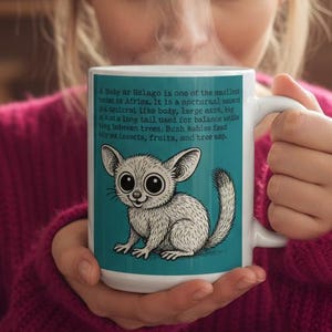 Könnte beinhalten: Weißer Keramikbecher mit türkisfarbenem Hintergrund, der eine Cartoon-Illustration eines kleinen Tieres mit großen Augen zeigt. Der Becher hat Text, der das Tier beschreibt. Die Person, die den Becher hält, trägt einen rosa Pullover.