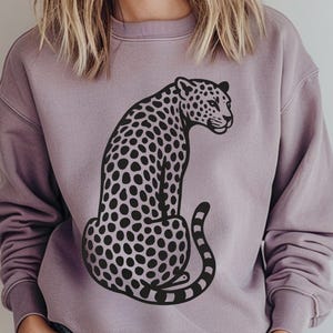 Pode incluir: Um moletom malva com um desenho de leopardo em preto e branco. O leopardo é retratado sentado, com a cauda enrolada. A palavra "Paragon" está impressa em escrita turquesa no canto superior direito do moletom.