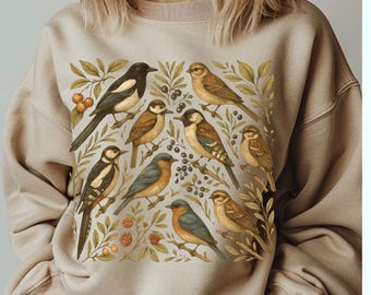 May include: Sand-colored sweatshirt featuring a detailed illustration of various birds amidst foliage and berries. The design includes birds in shades of brown, black, and blue, surrounded by green leaves and red berries. The word "Sand" is written in cursive at the top.