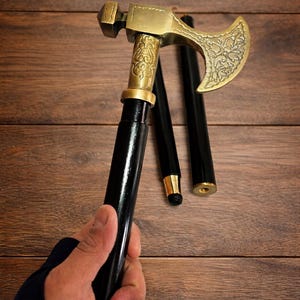 Puede incluir: Un bastón negro y dorado con una cabeza en forma de hacha. El bastón es sostenido por una mano, y la cabeza es de metal color oro con detalles ornamentados. El bastón está parcialmente desmontado, con las secciones a su lado.