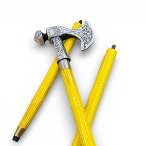 Może przedstawiać: Ozdobna laska do chodzenia ze srebrną głowicą topora i wzorem młotka. Rękojeść jest w jaskrawożółtym kolorze z pionowymi rowkami. Laska ma około 91 cm długości.