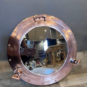 May include: A round, copper-colored porthole mirror with a polished finish. The mirror has a hinged top and two side latches. Rivets secure the frame. The mirror reflects the surrounding environment, including a person holding a phone.