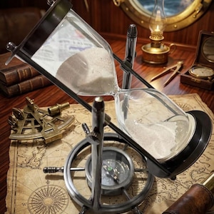 May include: A rotating hourglass with a metallic frame and white sand, mounted on a base shaped like a ship's wheel with an integrated compass. The hourglass is positioned on a vintage-style nautical map with a compass rose illustration.