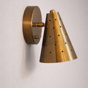 May include: A brass wall sconce with a conical shade and a circular backplate. The shade has small, evenly spaced holes. The fixture is mounted on a white wall, showcasing a vintage or mid-century modern design.