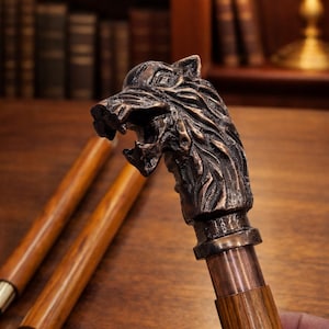 Puede incluir: Bastón con cabeza de lobo de bronce y mango de madera. La cabeza de lobo está detallada, con la boca abierta y dientes visibles. El bastón se muestra sobre una superficie de madera, sugiriendo un estilo clásico o vintage.