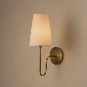 Op de afbeelding: Een wandlamp met een conische, gebroken witte stoffen kap bevestigd aan een gebogen arm van geborsteld messing. De armatuur is voorzien van een ronde messing muurplaat die de arm met de muur verbindt voor een eenvoudig ontwerp.