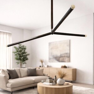 May include: Contemporary black and brass chandelier with three horizontal arms and a central vertical support. The light fixture has a minimalist design. The chandelier is suspended from the ceiling in a living room setting.