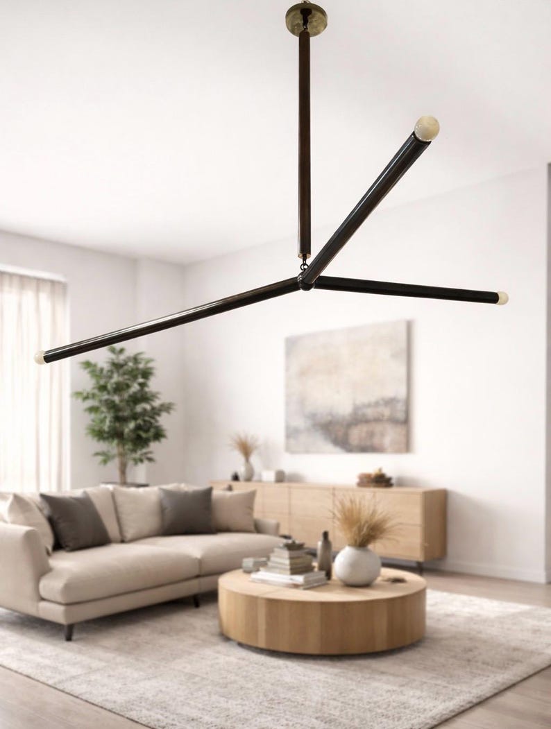 May include: Contemporary black and brass chandelier with three horizontal arms and a central vertical support. The light fixture has a minimalist design. The chandelier is suspended from the ceiling in a living room setting.