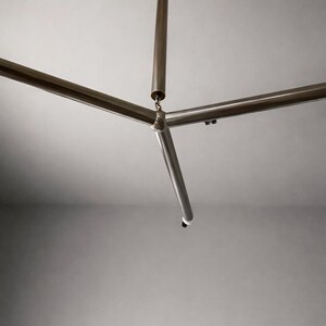 May include: A contemporary ceiling light fixture featuring a three-arm design. The light has a central vertical rod and three horizontal rods extending outwards. The metal finish is a brushed brass colour, and the fixture is mounted on a white ceiling.