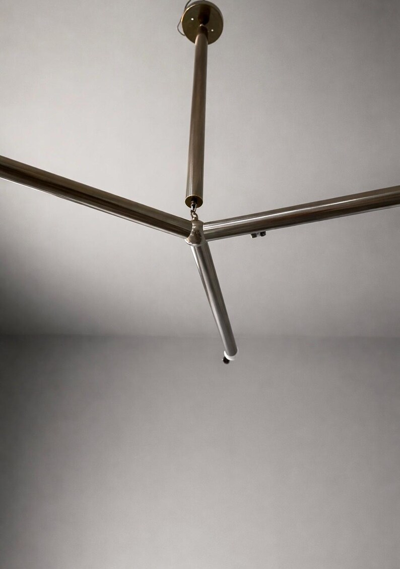 May include: A contemporary ceiling light fixture featuring a three-arm design. The light has a central vertical rod and three horizontal rods extending outwards. The metal finish is a brushed brass colour, and the fixture is mounted on a white ceiling.