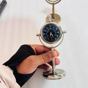 May include: A silver-colored desk clock with a dark blue face and white numerals. The clock has a round face with a silver frame and a stand. Another similar clock is in the background. The clock is being held by a person.