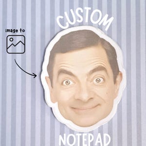 以下が含まれることがあります： 笑顔の顔が特徴のカスタムメモ帳。上部に「CUSTOM」、下部に「NOTEPAD」の文字。背景は青と白の縦縞で、左上には画像アイコンと矢印があります。