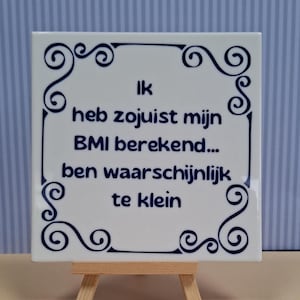 Può includere: Una piastrella di ceramica bianca con un bordo decorativo blu e testo in olandese. Il testo recita: "Ik heb zojuist mijn BMI berekend... ben waarschijnlijk te klein." La piastrella è esposta su un piccolo cavalletto di legno.