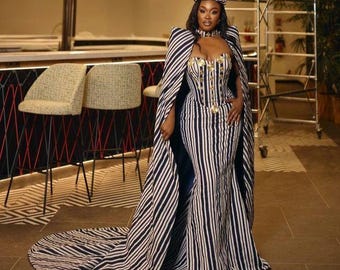 May include: A woman wearing a black and white striped dress with a matching cape. The dress has a fitted bodice and a flowing skirt. The woman is wearing a gold headpiece and has her hair pulled back.