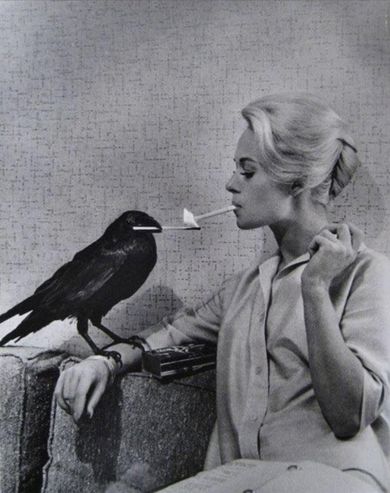 May include: Black and white photograph of a woman smoking a cigarette lit by a raven. The raven is perched on the arm of a sofa, holding a lit match in its beak. The woman is wearing a button-down shirt.