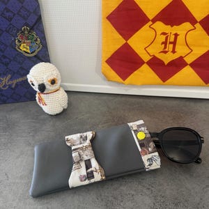 Op de afbeelding: Grijze zonnebril etui met een patroonstof accent en een gele knoopsluiting. Zwarte zonnebril rust op de etui. Een witte gehaakte uil en een Hogwarts-thema banner staan op de achtergrond.