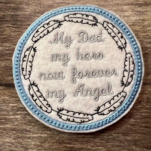 May include: A white felt circle with a blue embroidered border. The circle has black embroidered feathers around the edge and the text "My Dad my hero now forever my Angel" in silver thread.