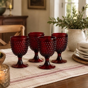 May include: A set of four deep red glass goblets featuring a textured hobnail pattern on the exterior. Each goblet has a short, sturdy stem and a wide circular base, crafted from translucent red glass that catches the light.