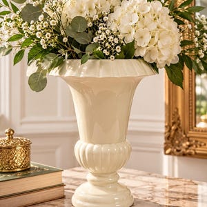 Puede incluir: Un florero de pedestal de cerámica color crema con un borde ancho y acanalado y una sección central redondeada y estriada, lleno de un gran arreglo de flores de hortensia blancas, paniculata y hojas de eucalipto verdes.