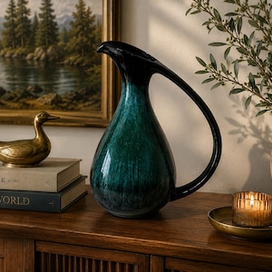 May include: A ceramic pitcher with a teardrop-shaped body featuring a deep teal and black gradient glaze. The pitcher has a long, curved handle extending from the top rim to the base and a pointed spout for pouring, showcasing a smooth, glossy finish.