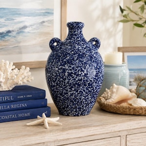 Puede incluir: Un jarrón de cerámica azul marino moteado con dos asas. El jarrón se exhibe sobre una superficie de madera clara con decoración costera, que incluye libros, una escultura de coral y conchas marinas. Una pintura de paisaje marino enmarcada está en el fondo.