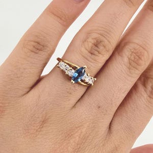 Puede incluir: Un anillo dorado con una piedra preciosa azul oscuro en forma de pera como pieza central. El anillo está adornado con piedras preciosas más pequeñas y transparentes y tiene un diseño de banda dividida. El anillo está en un dedo.