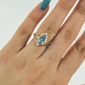 Puede incluir: Un anillo de oro con una piedra preciosa azul claro en forma de marquesa, flanqueada por piedras claras más pequeñas. El anillo tiene un diseño de banda retorcida. El anillo se muestra en una mano con esmalte de uñas verde azulado y rosa.