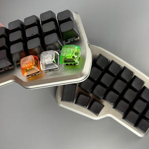 May include: Ergonomic split keyboard with black keycaps and a silver frame. Three colorful keycaps in orange, clear, and green are visible. The keyboard is designed for comfort and efficiency.