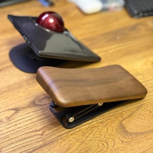 May include: A black trackball mouse with a red ball and a wooden wrist rest are displayed on a wooden surface. The trackball has a triangular design. The wrist rest is made of dark wood and has a curved shape.