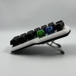 May include: A compact, white and black mechanical keyboard with a variety of keycap colors. The keyboard is angled upward with a silver stand. The keycaps include black, green, blue, and brown colors.