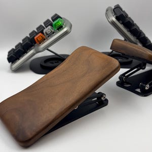 May include: Two ergonomic palm rests featuring smooth, dark brown wooden surfaces mounted on adjustable black metal scissor-lift bases, positioned in front of two mechanical keypads with black keys and visible orange and green switches.