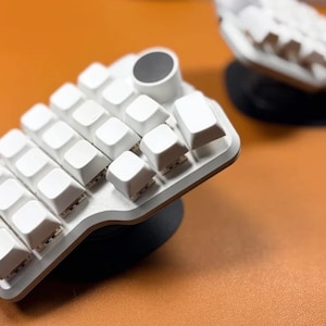 Könnte beinhalten: Eine weiße ergonomische Tastatur mit geschwungenem Design und weißen Tastenkappen. Die Tastatur hat eine kreisförmige Auflage und befindet sich vor einem orangefarbenen Hintergrund. Eine weitere Tastatur ist im Hintergrund sichtbar.
