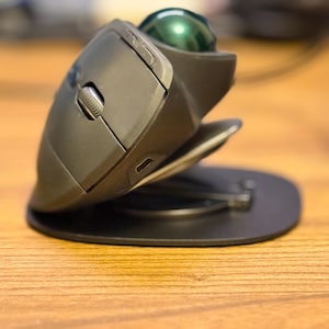 Könnte beinhalten: Eine schwarze ergonomische Trackball-Maus mit einem grünen Trackball und einem silbernen Scrollrad. Die Maus liegt auf einer schwarzen Basis auf einer Holzoberfläche.