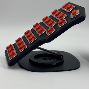 Peut inclure: Un testeur d'interrupteurs de clavier mécanique noir avec des interrupteurs rouges. Le testeur est incliné vers le haut et soutenu par une base noire. Les interrupteurs sont disposés en rangées et en colonnes.