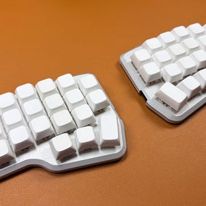 May include: A split ergonomic mechanical keyboard featuring two separate sections with white blank keycaps on a silver-colored base. The layout is non-staggered with a contoured shape, designed for two-handed use with a distinct thumb cluster area.