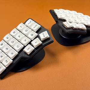May include: A split ergonomic keyboard featuring two black contoured bases with white keycaps. Each half has a staggered layout with letters, numbers, and function keys including Tab, Ctrl, Shift, Cmd, Space, Pg Up, and Pg Dn printed in black.