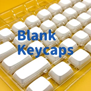 Puede incluir: Una bandeja de plástico transparente llena de teclas blancas en blanco. La bandeja está sobre una superficie amarilla. El texto "Blank Keycaps" está escrito en letras azules en la bandeja.